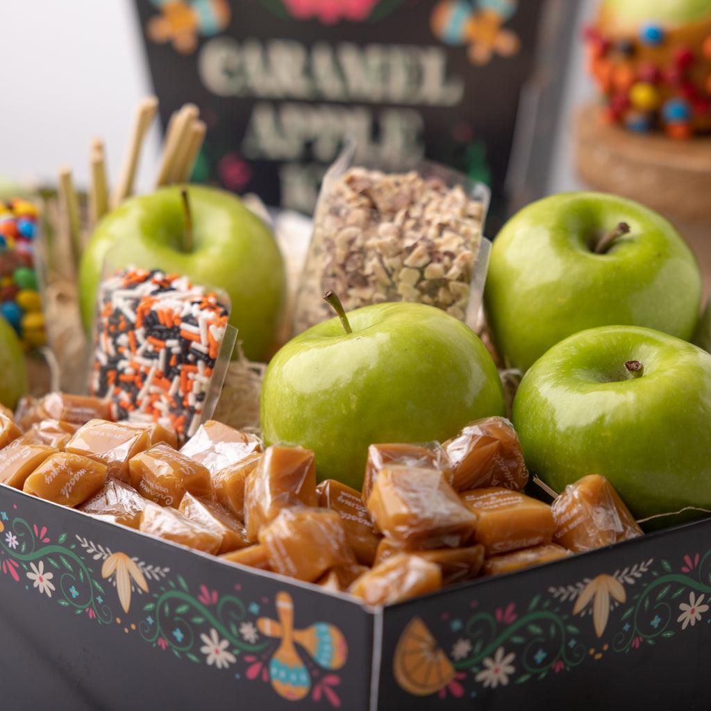 Recipe card for the Caramel Apple Kit A person holding the Caramel Apple Kit  Apple, candies, and caramels in a festive box Coffin shaped caramel apple kit for dia de muertos Caramel apples with M&M's and nuts M&M's and green apples in a festive box Caramels, candies, and green apples in a festive box Bags of candy in the Dia de Muertos box Openned transparent bag of M&M's