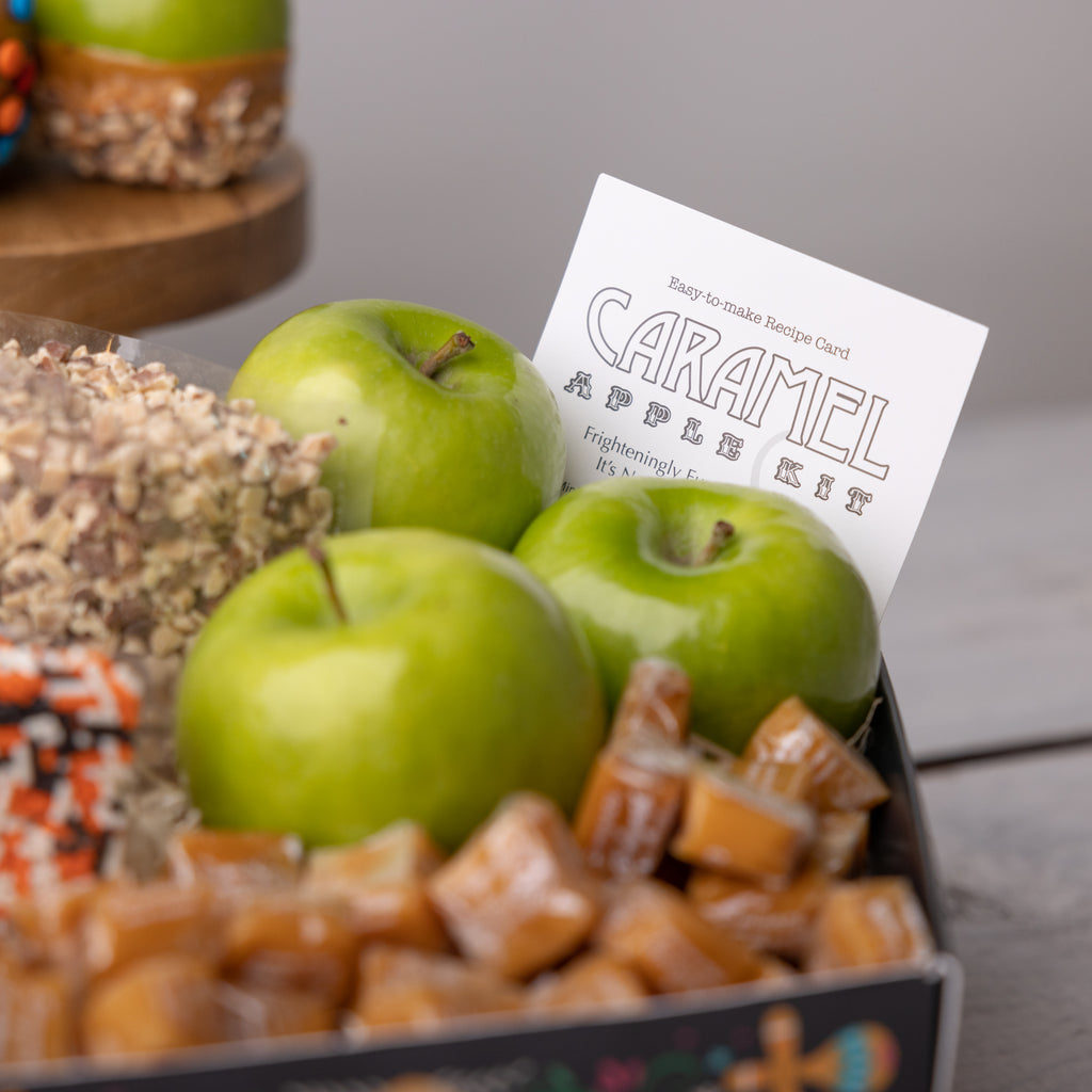 Recipe card for the Caramel Apple Kit A person holding the Caramel Apple Kit  Apple, candies, and caramels in a festive box Coffin shaped caramel apple kit for dia de muertos Caramel apples with M&M's and nuts M&M's and green apples in a festive box Caramels, candies, and green apples in a festive box Bags of candy in the Dia de Muertos box Openned transparent bag of M&M's